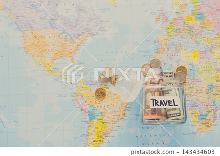 A travel savings jar sits on a world map. Coins and bills are scattered around the jar. Different countries are visible on the map. The map shows regions in North and South America. A travel savings jar sits on a world map. Coins and bills are scattered around the jar. Different countries are visible on the map. The map shows regions in North and South America. 143434603