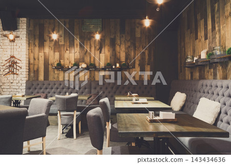 Seating arrangement in a restaurant featuring wooden walls, tables, and chairs. The space is well lit with warm lights and has a welcoming atmosphere for guests. Seating arrangement in a restaurant featuring wooden walls, tables, and chairs. The space is well lit with warm lights and has a welcoming atmosphere for guests. 143434636