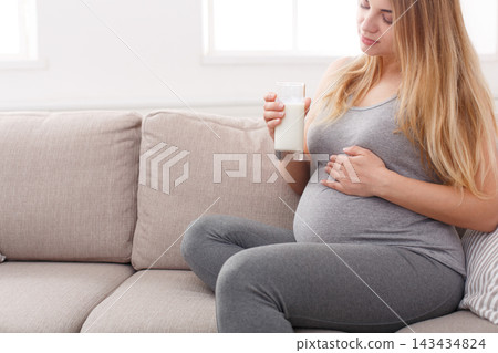 A pregnant woman sits on a couch in a living room, holding a glass of milk. She gently rests her hand on her belly, enjoying a moment of relaxation. A pregnant woman sits on a couch in a living room, holding a glass of milk. She gently rests her hand on her belly, enjoying a moment of relaxation. 143434824
