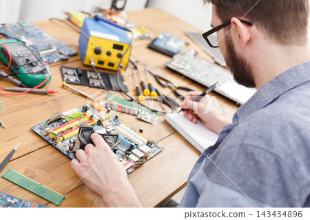 A man sits at a wooden table working on a computer motherboard. Tools like screwdrivers and a soldering iron are nearby. He takes notes in a notebook as he focuses on the task. A man sits at a wooden table working on a computer motherboard. Tools like screwdrivers and a soldering iron are nearby. He takes notes in a notebook as he focuses on the task. 143434896