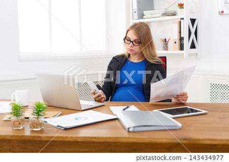 A pregnant woman sits at a wooden desk in a bright office. She holds a phone in one hand and papers in the other, focused on her work. The laptop is open and small plants are on the desk. A pregnant woman sits at a wooden desk in a bright office. She holds a phone in one hand and papers in the other, focused on her work. The laptop is open and small plants are on the desk. 143434977