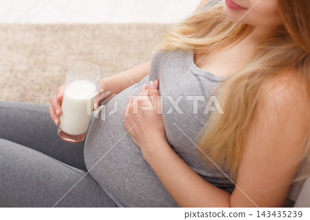 A woman sits on the floor with her legs extended. She holds a glass of milk in one hand and gently rests her other hand on her pregnant belly. The setting appears cozy and warm. 143435239