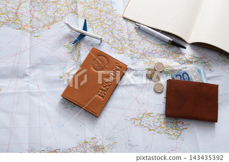 A workspace shows a passport, coins, and cash placed on a map. An airplane model and a pen are also on the table. This scene suggests travel planning and preparation. 143435392