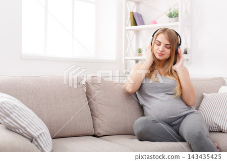 A woman who is pregnant sits on a couch in a bright room. She listens to music with headphones on and enjoys her time. Sunlight comes through the window, creating a warm atmosphere. A woman who is pregnant sits on a couch in a bright room. She listens to music with headphones on and enjoys her time. Sunlight comes through the window, creating a warm atmosphere. 143435425