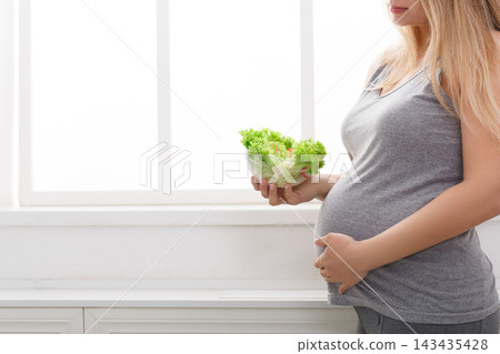 A pregnant woman stands by a window holding a bowl of fresh lettuce. She is in a light room with white walls. 143435428