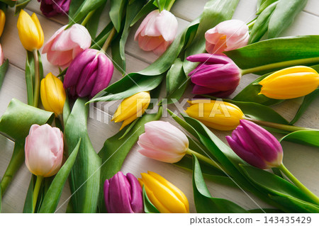 Different shades of tulips lay on a wooden table. The flowers include pink, yellow, and purple tulips with green leaves. The arrangement shows a mix of colors without any specific pattern. 143435429