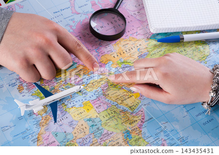 Two hands point at different locations on a map while planning a trip. A magnifying glass is nearby, along with a notepad and pens. An airplane model is placed on the map. 143435431