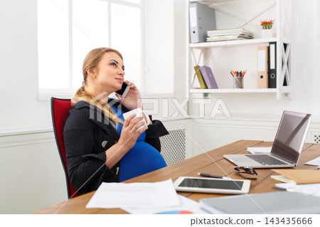A pregnant woman is sitting at a desk in her home office. She is on the phone and holding a cup. Papers and a laptop are on the desk. The room has natural light from a window. 143435666