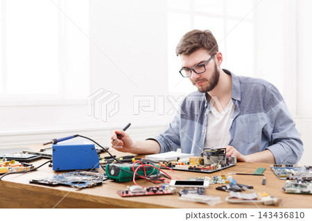 A young man works on repairing various electronic devices on a wooden table. Tools and components are spread around him in a well-lit room, showing a hands-on activity with attention to detail. 143436810