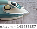Modern steam iron pressing a blue shirt on a bed, representing household laundry, clothing care and wrinkle removal. 143436857