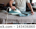 Woman using a steam iron to press a blue shirt on a bed, illustrating household chores, garment care and everyday home life 143436860
