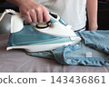 Woman ironing a blue shirt on a bed with a steam iron, representing everyday housework, clothing care and domestic routine. 143436861