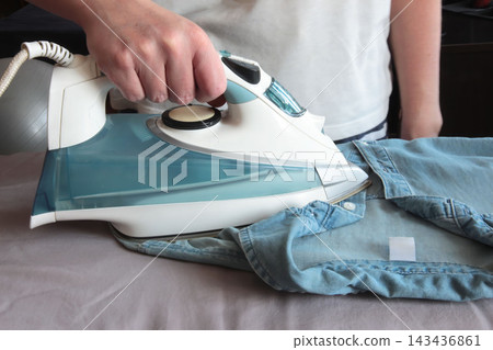 Woman ironing a blue shirt on a bed with a steam iron, representing everyday housework, clothing care and domestic routine. Woman ironing a blue shirt on a bed with a steam iron, representing everyday housework, clothing care and domestic routine. 143436861