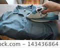Person ironing a blue shirt with a steam iron at home, illustrating clothing care, household chores and garment maintenance. 143436864
