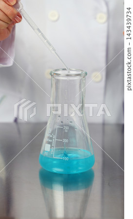 Scientist's hand using a dropper to add blue liquid into a conical flask during a chemistry experiment in a laboratory Scientist's hand using a dropper to add blue liquid into a conical flask during a chemistry experiment in a laboratory 143439734