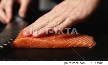 Hands of professional chef slicing fresh raw salmon fillet for sushi preparation on a dark cutting board Hands of professional chef slicing fresh raw salmon fillet for sushi preparation on a dark cutting board 143439849