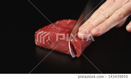 Hand slicing fresh raw tuna steak with a sharp knife on a dark background for gourmet seafood preparation Hand slicing fresh raw tuna steak with a sharp knife on a dark background for gourmet seafood preparation 143439850
