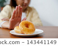 glazed donuts on plate with Asian woman making stop hand gesture in background, refusing unhealthy sugary food for diet and weight loss healthy concept. 143441149