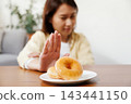 glazed donuts on plate with Asian woman making stop hand gesture in background, refusing unhealthy sugary food for diet and weight loss healthy concept. 143441150