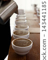 barista or q grader performing coffee cupping to evaluate aroma and flavor quality, professional coffee tasting process with multiple cups on wooden table during specialty coffee quality control 143441185
