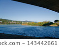 View from under a concrete bridge across a calm blue river to green hills 143442661
