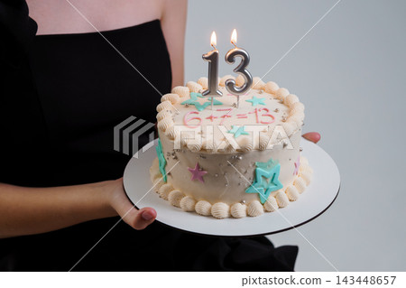 Woman in black dress holding a cream birthday cake with number 13 candles and colorful star decorations Woman in black dress holding a cream birthday cake with number 13 candles and colorful star decorations 143448657