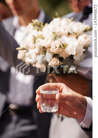 Groom holds wedding toast glass with bride bouquet in background celebrating marriage ceremony outdoors sunny day Groom holds wedding toast glass with bride bouquet in background celebrating marriage ceremony outdoors sunny day 143448673