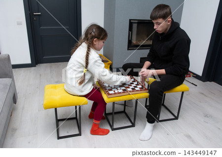 In the warm and inviting atmosphere of their cozy living room, a captivating chess match unfolds between siblings, filling the afternoon with strategic moves. 143449147