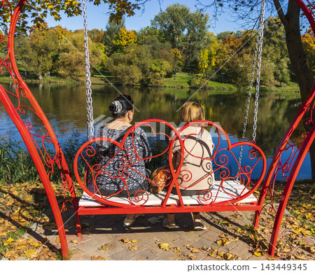 Minsk, Belarus - October 5, 2026: Two women sitting on a red heart shaped swing by a pond Minsk, Belarus - October 5, 2026: Two women sitting on a red heart shaped swing by a pond 143449345