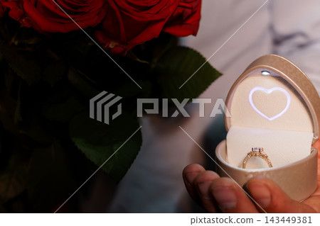Close up of hand holding open ring box with heart light next to red roses proposal scene Close up of hand holding open ring box with heart light next to red roses proposal scene 143449381