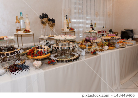 Elegant buffet table with fresh fruit tarts chocolate mousse cakes wine bottles white tablecloth event catering Elegant buffet table with fresh fruit tarts chocolate mousse cakes wine bottles white tablecloth event catering 143449406