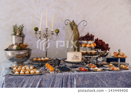 Elegant dessert table spread featuring small cakes, fresh fruit, and silver candelabra with white candles and purple velvet cloth Elegant dessert table spread featuring small cakes, fresh fruit, and silver candelabra with white candles and purple velvet cloth 143449408