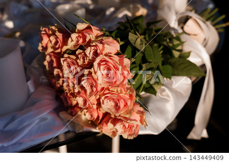 Bouquet of peach roses with white satin ribbon lying on a table in warm sunlight and green leaves Bouquet of peach roses with white satin ribbon lying on a table in warm sunlight and green leaves 143449409