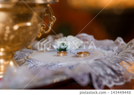 Gold wedding rings on white satin with lace and a silver chalice in warm church lighting Gold wedding rings on white satin with lace and a silver chalice in warm church lighting 143449448