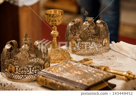 Gilded Orthodox Royal Crowns and Gold Chalice Resting on White Lace Tablecloth in Church Setting with Intricate Filigree 143449452