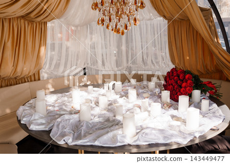 Romantic dinner table with white candles red roses gold drapes chandelier luxury hotel setup for date night proposal or anniversary celebration Romantic dinner table with white candles red roses gold drapes chandelier luxury hotel setup for date night proposal or anniversary celebration 143449477