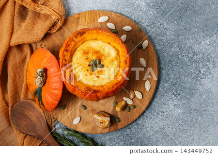 Pumpkin cream soup served in a hollowed pumpkin with pumpkin seeds and garlic on a wooden board at a kitchen table Pumpkin cream soup served in a hollowed pumpkin with pumpkin seeds and garlic on a wooden board at a kitchen table 143449752