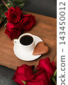 Red roses surround a coffee cup and chocolate on a wooden tray with a dark background 143450012