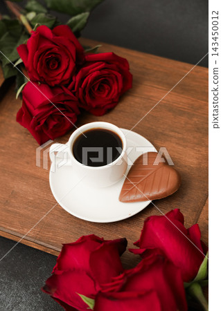 Red roses surround a coffee cup and chocolate on a wooden tray with a dark background Red roses surround a coffee cup and chocolate on a wooden tray with a dark background 143450012