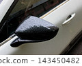 The black side mirror of a white car is covered with large raindrops 143450482