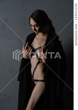 Woman in black leather harness and open coat against dark textured wall, dramatic side lighting, moody atmosphere Woman in black leather harness and open coat against dark textured wall, dramatic side lighting, moody atmosphere 143451238