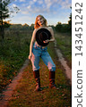 Woman in jeans and tall boots holding a black hat on a rural path 143451242