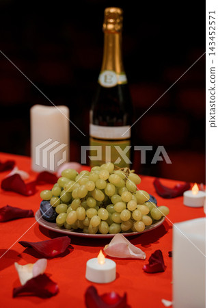 Green grapes on a plate with wine bottle and tealights on red tablecloth featuring scattered rose petals in romantic setting Green grapes on a plate with wine bottle and tealights on red tablecloth featuring scattered rose petals in romantic setting 143452571