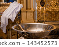 Silver baptismal font with three lit candles and white lace cloth in ornate golden church interior 143452573