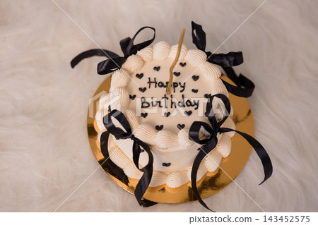 White birthday cake with black ribbons and candles on a gold plate, elegant party dessert with happy message in hearts. 143452575