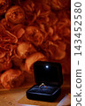 Gold diamond ring in open black velvet box surrounded by warm blurred orange roses for romantic proposal context 143452580