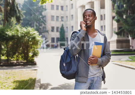 A student stands on a college campus speaking on the phone. He carries a backpack and holds books in his arms. The background shows a building and trees under clear skies during the day. A student stands on a college campus speaking on the phone. He carries a backpack and holds books in his arms. The background shows a building and trees under clear skies during the day. 143453801