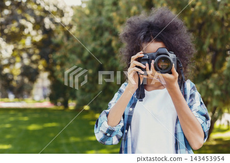A woman stands in a park holding a camera and looking through the lens. Trees surround her while she prepares to take a photo. Sunlight shines through the leaves. 143453954