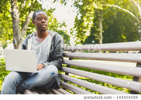A young man sits on a bench in a park while using a laptop. He looks thoughtful and relaxed. The sun shines through the trees creating a pleasant setting. 143454389