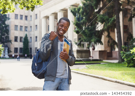 A student stands on a walkway at school, holding books and looking happy. Other students walk in the background. The buildings are tall and the trees are green. It is daytime. A student stands on a walkway at school, holding books and looking happy. Other students walk in the background. The buildings are tall and the trees are green. It is daytime. 143454392